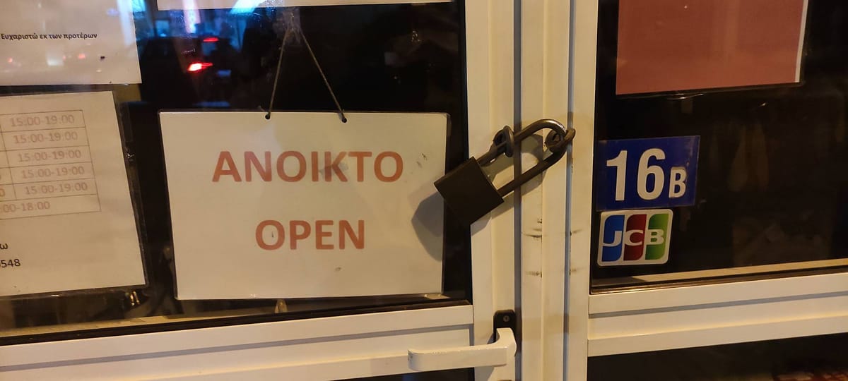 Auf dem Schild an der Tür eines Ladengeschäfts steht "ANOIKTO - OPEN", während gleichzeitig ein großes Schloss vor der Tür hängt.